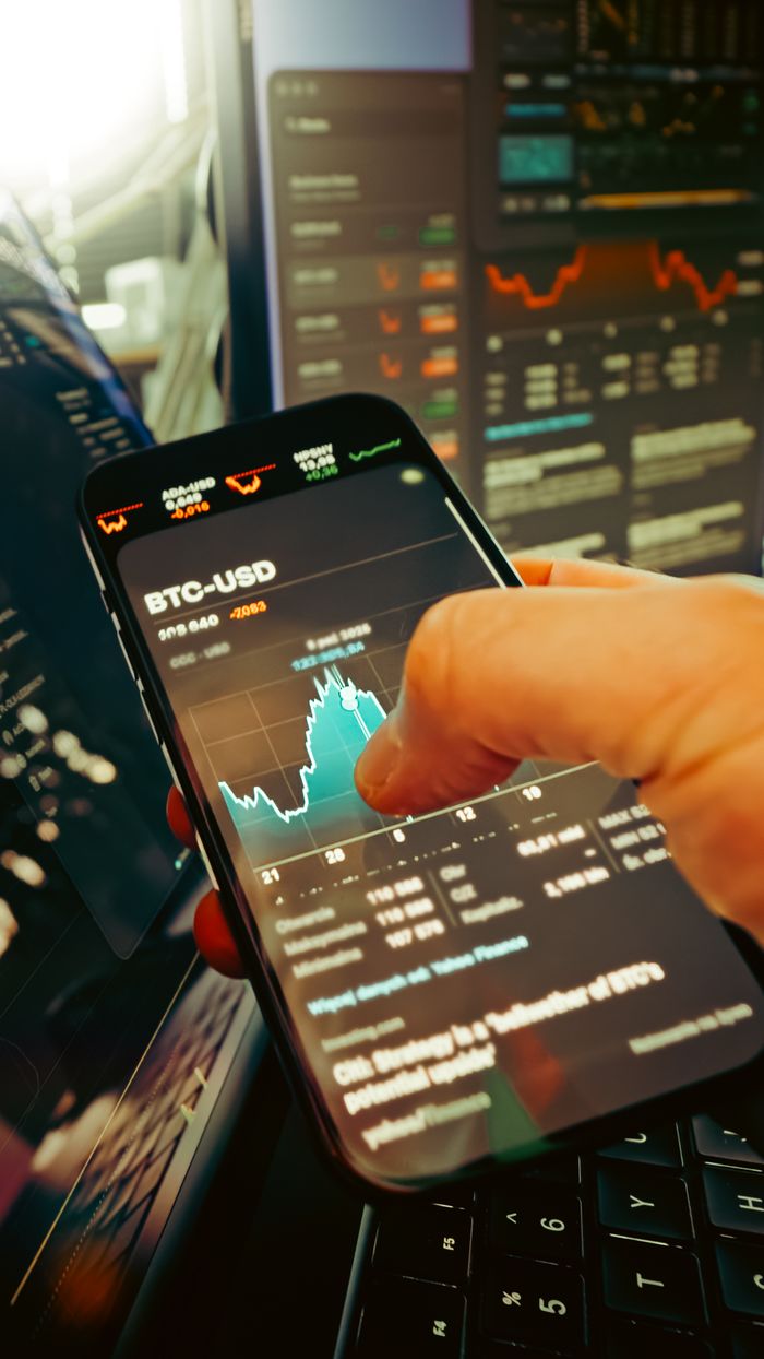 Une main tient un smartphone affichant le graphique BTC-USD au milieu d’écrans de trading numériques, soulignant la finance crypto.